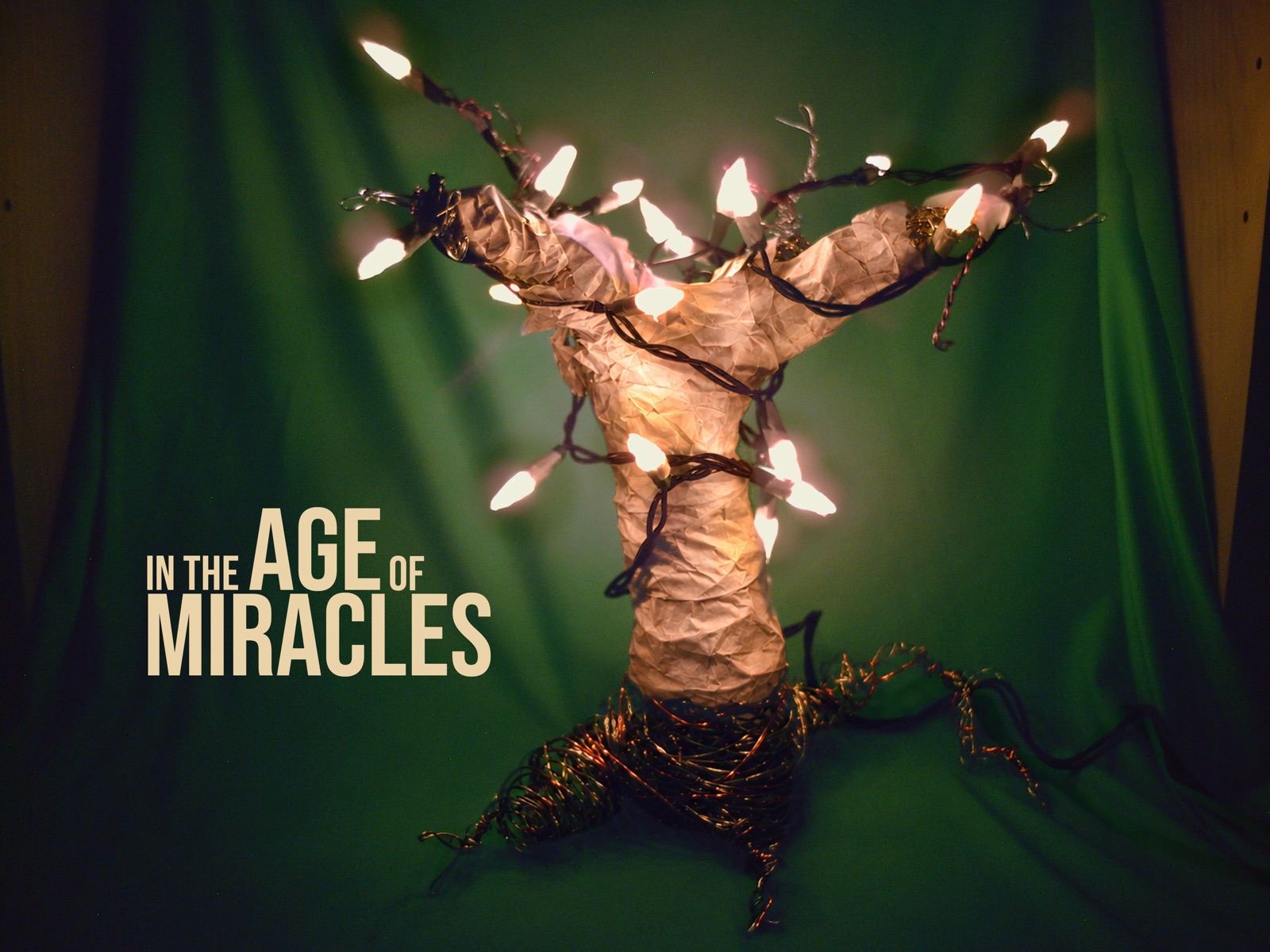 A handmade branching sculpture wrapped in glowing pink lights against deep green fabric, with warm gold In the Age of Miracles lettering.